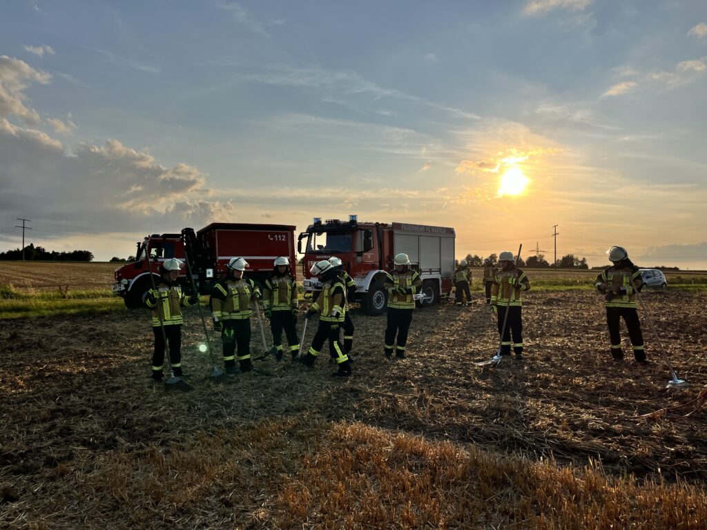 mehrere feuerwehrangehörige mit schutzkleidung vor feuerwehrfahrzeugen auf einem feld im sonnenuntergang